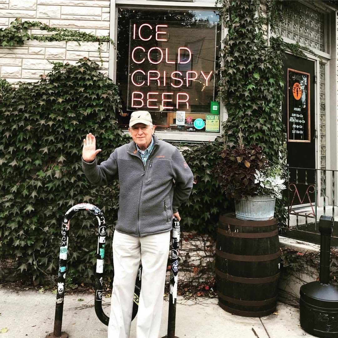 Dan Pelzer waving outside a Columbus storefront beneath an ‘ICE COLD CRISPY BEER’ neon sign